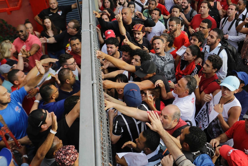 People block an entrance and seem agitated. Fence in the middle of the picture.