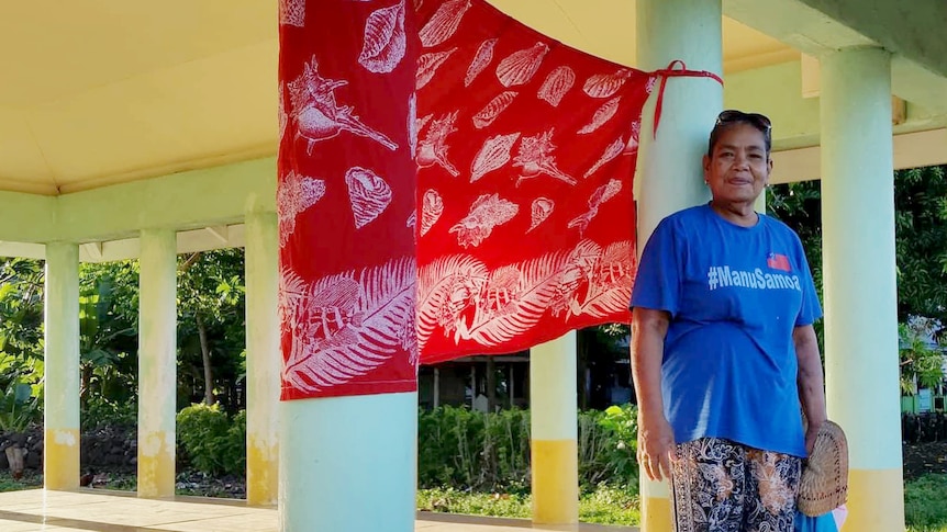 A woman in Samoa stands beside a red flag, put up near the front of a property to let medical teams know they need vaccinations.