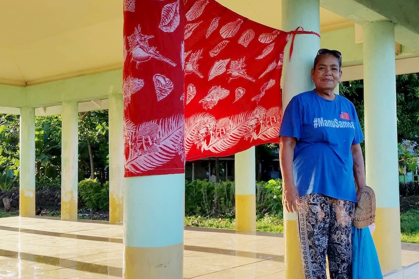 A woman in Samoa stands beside a red flag, put up near the front of a property to let medical teams know they need vaccinations.