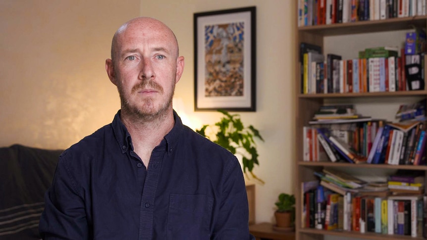 A man sitting indoors, behind him is a bookshelf and a potted plant.