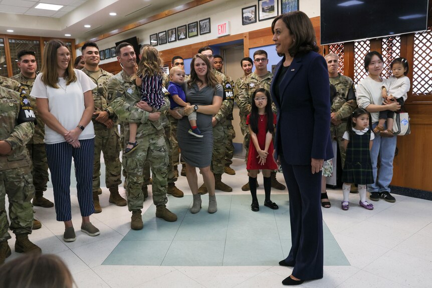 Kamala Harris stands in front of a group of American soldiers and their wives and children. 