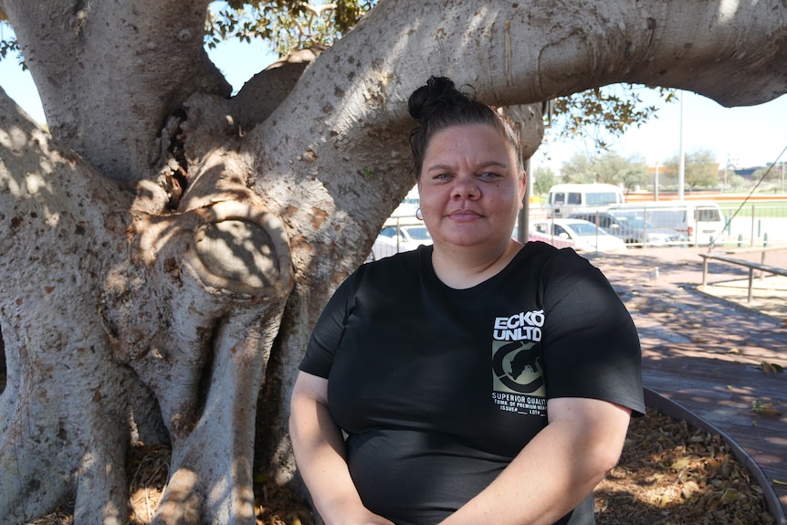 A woman stands in under a large tree facing the camera in a black t-shit, hands clapsed in front.