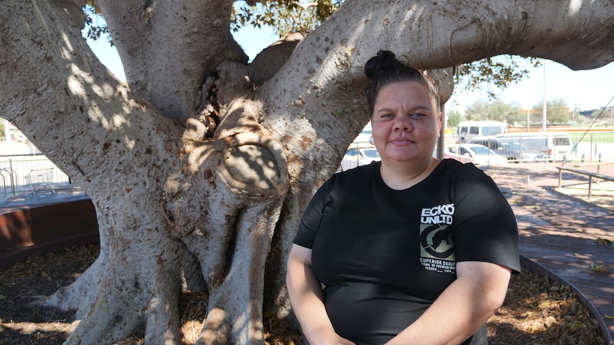 A woman stands in under a large tree facing the camera in a black t-shit, hands clapsed in front.