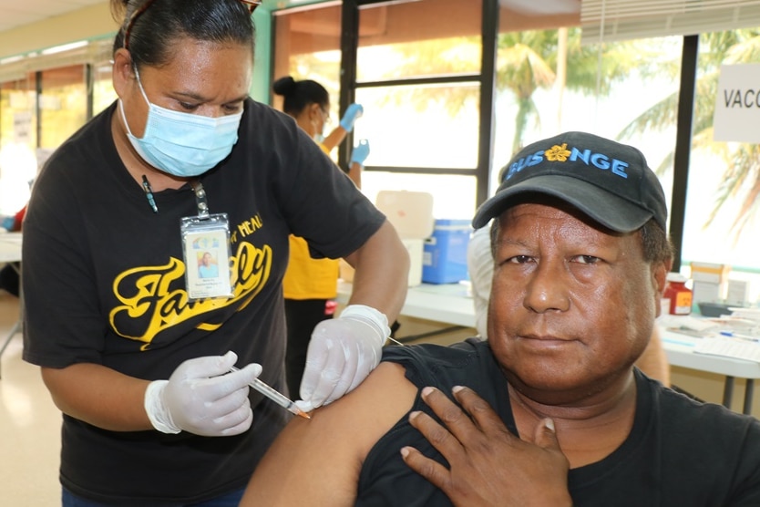 You view a man in a black short-sleeve t-shirt looking at the camera as a woman in a mask pushes a needle into his right arm.