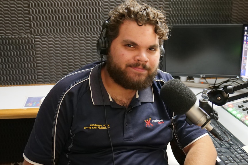 A young, dark-bearded Aboriginal man behind a microphone in an outback radio studio