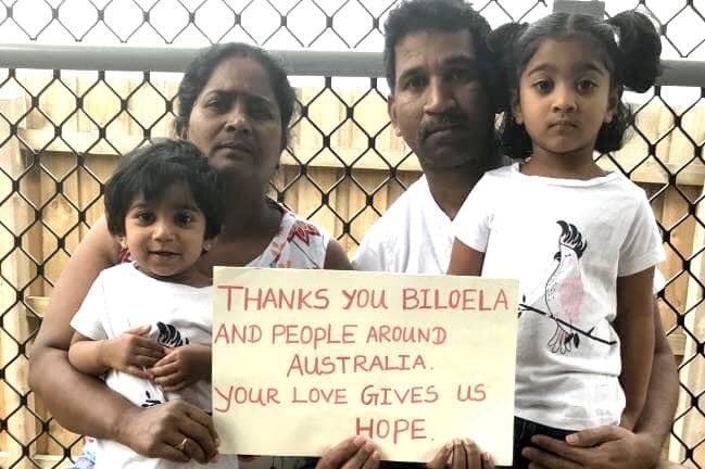 Family sit against a fence holding a sign that reads: "Thanks you Biloela and people around Australia. You give us hope".