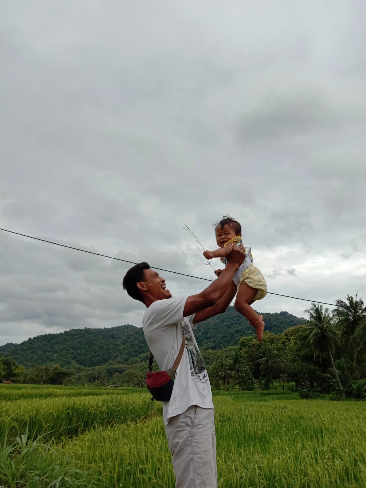 A man lifts a daughter about two years old against the backdrop of rice fields, both of them laughing.