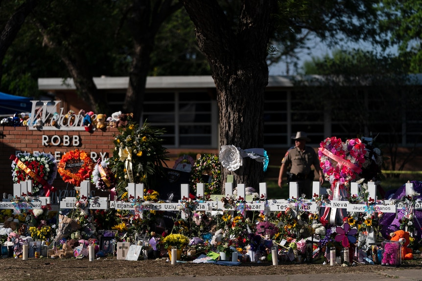 Cruces blancas y muchas flores rodean el letrero de la escuela en Robb Elementary después de una masacre, con la policía al fondo
