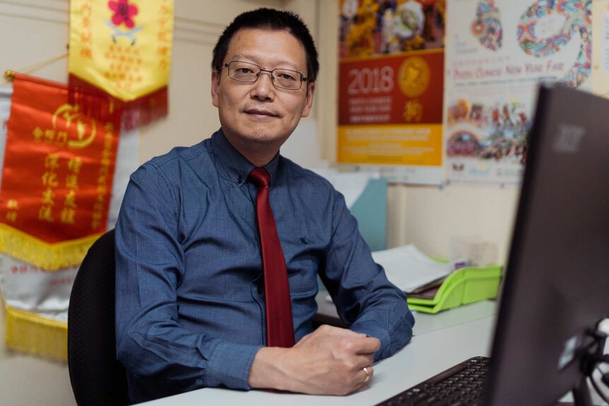 A man in a blue shirt and red tie sits at a desk with Chinese banners on the walls.