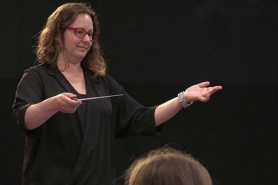 Photograph of a middle-aged woman with red glasses conducting musicians