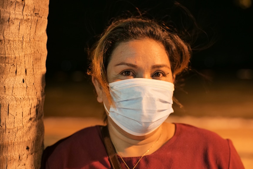 A woman wearing a white mask and red top stares at the camera while standing next to a tree.