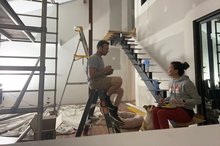 Tamiko Gleeson and her husband eating dinner in their living room while it's renovated. A scaffold is visible on the left