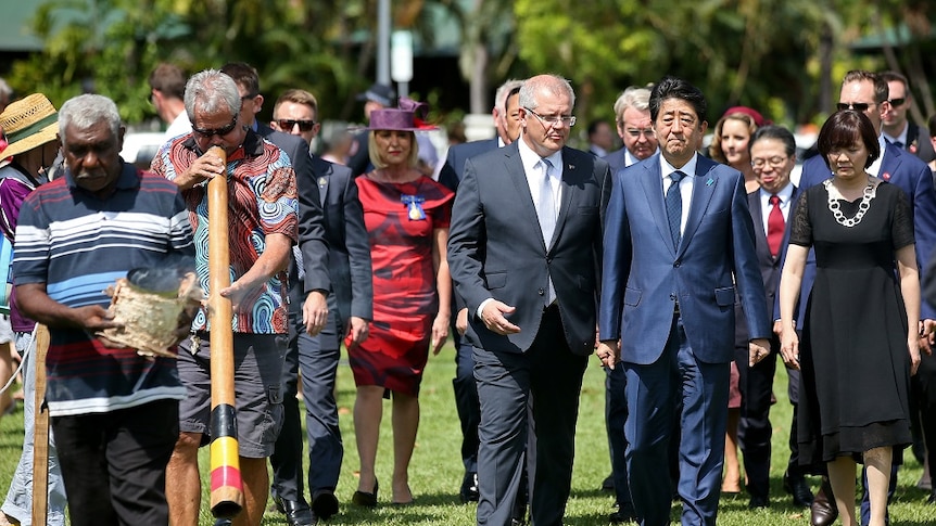 Shinzo Abe and Scott Morrison walk beside Aboriginal men playing the didgeridoo and carrying a ceremonial fire
