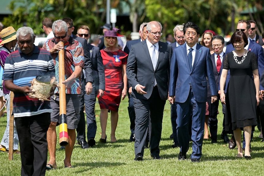 Shinzo Abe and Scott Morrison walk beside Aboriginal men playing the didgeridoo and carrying a ceremonial fire