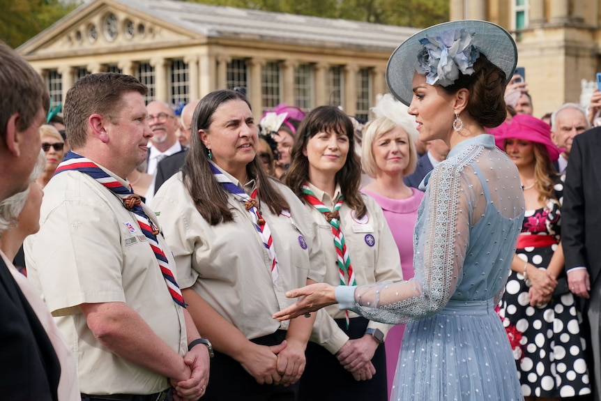 A woman wearing a blue dress and fascinator is speaking to three people in scouting uniforms while spectators look on.
