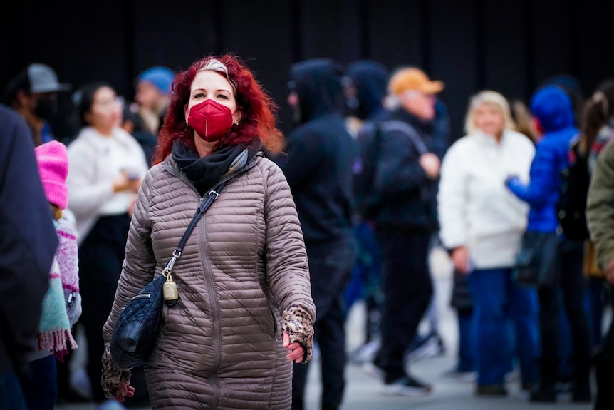 A woman with bright red hair, dressed in a red KN95 and beige puffer coat walks through a crowd