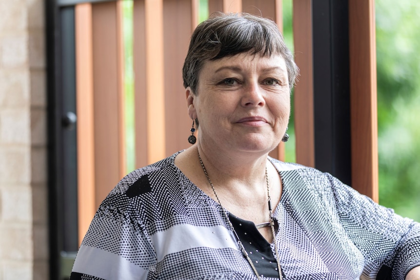 A woman with short brown hair wearing earrings and a grey patterned shirt smiles.