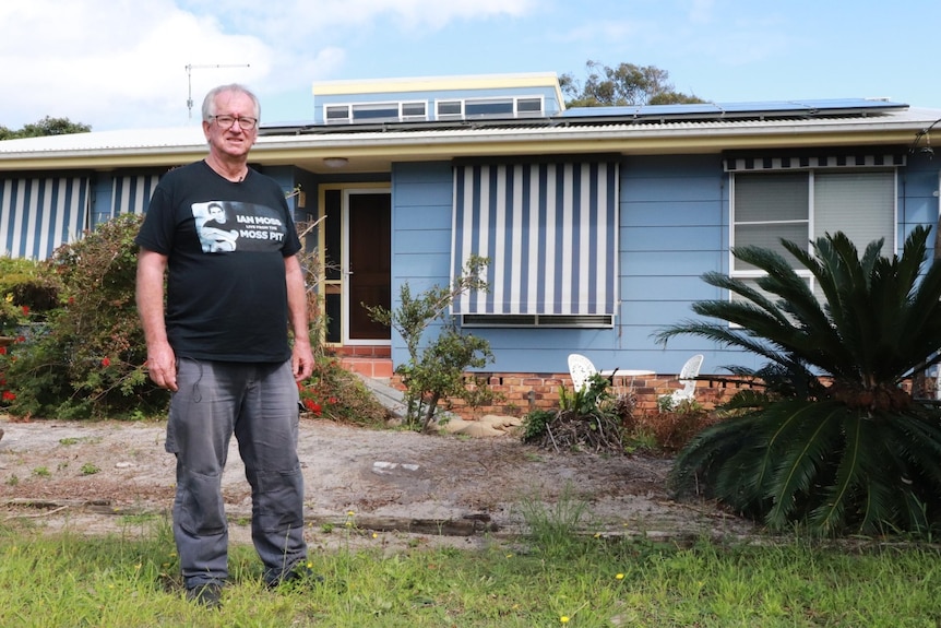 A man with grey hair, wearing a black t-shirt and grey pants and a glasses stands in front of a blue weatherboard.