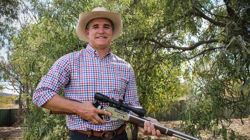 Robbie Katter holds a rifle at his rural property, surrounded by trees.