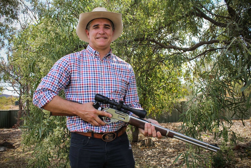 Robbie Katter holds a rifle at his rural property, surrounded by trees.