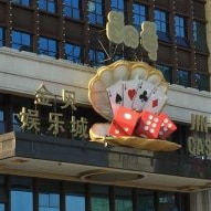 A high-rise building with a deck of cards and dice in an open oyster with signs Jin Bei Casino in English and Mandarin.