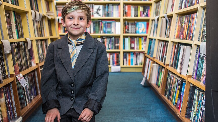 Colour photograph of a young boy wearing a tie and suit jacket, amongst book shelves in a bookshop.