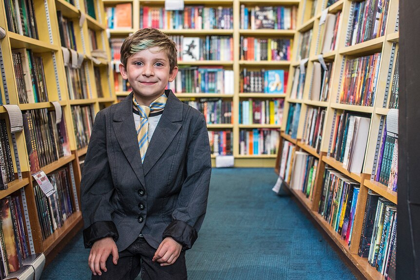 Colour photograph of a young boy wearing a tie and suit jacket, amongst book shelves in a bookshop.