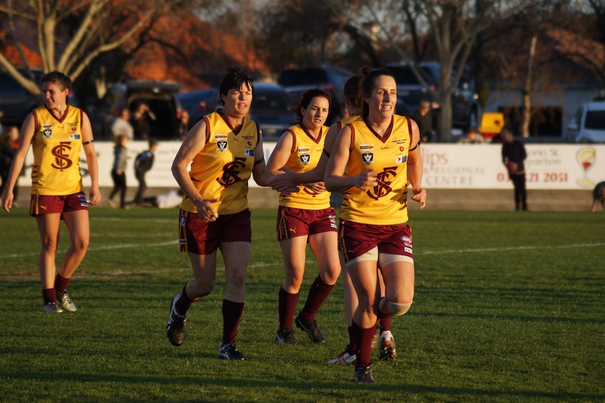 Women in gold and maroon football uniforms jog off the field