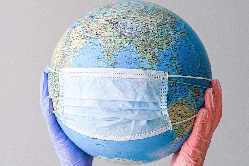 A globe wearing a mask, held by two gloved hands, the COVID-19 pandemic is affecting the world in 2021.