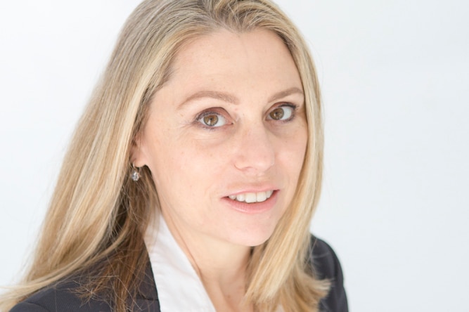 A woman in a grey suit and white shirt looks at the camera.