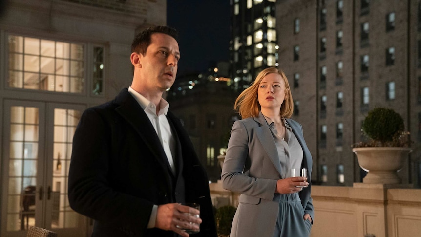 The two actors, both wearing suits, stand on a inner-city rooftop holding glasses of scotch.