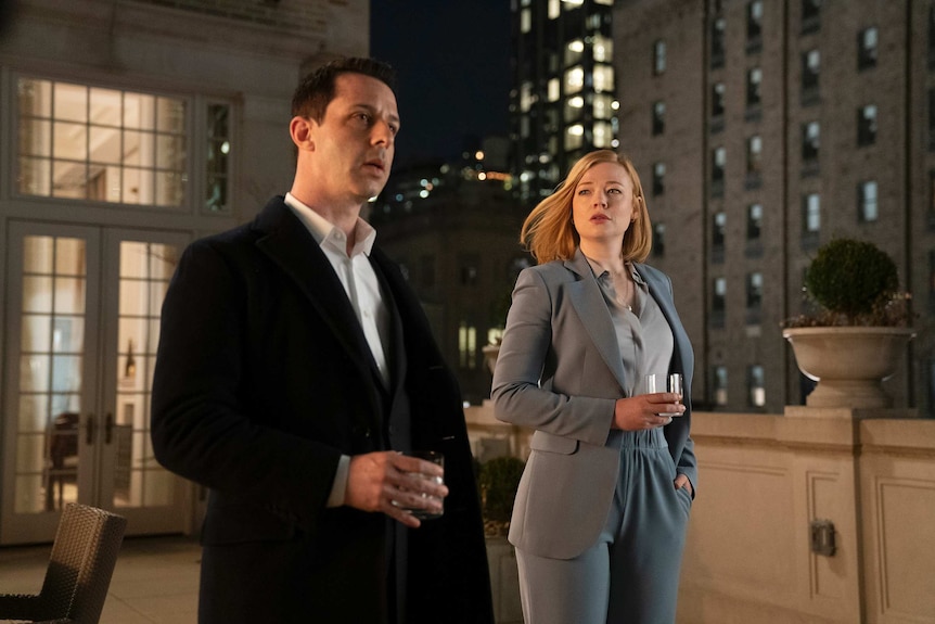 The two actors, both wearing suits, stand on a inner-city rooftop holding glasses of scotch.