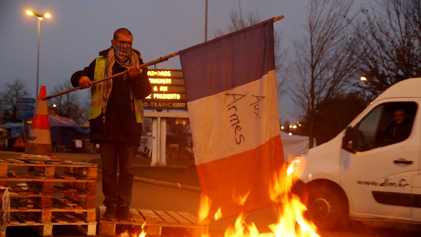 A man wearing a scarf over his nose and mouth holds a French flag near a pile of burning debris.