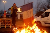 A man wearing a scarf over his nose and mouth holds a French flag near a pile of burning debris.