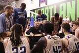 A basketball coach crouches down and gestures in a team talk to his players during a break in an NBL game.