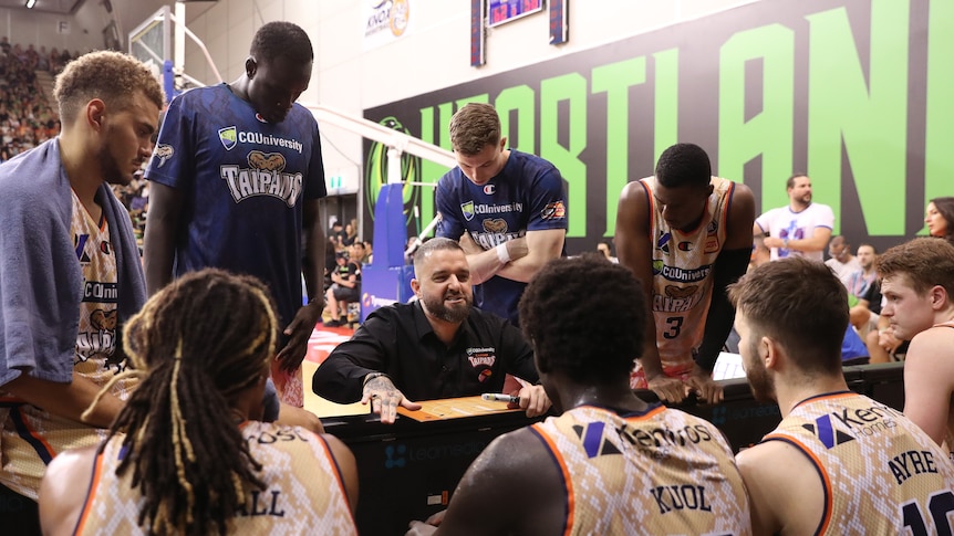 A basketball coach crouches down and gestures in a team talk to his players during a break in an NBL game.