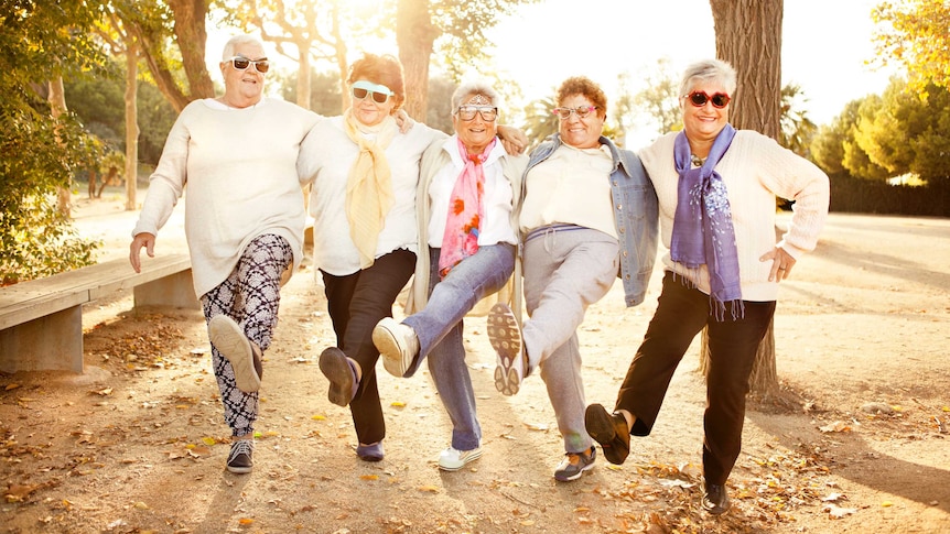 Five women wearing novelty sunglasses link arms and each kick a leg out as if dancing, while smiling or laughing.