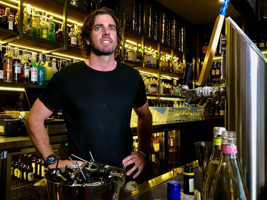 Glen stands with hands on hips behind the bar, rows of alcohol behind him
