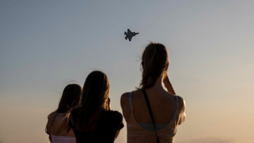 A silhouette of three young women or girls in front of a dusky sky with a fighter jet in the middle of it.