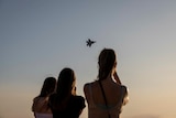 A silhouette of three young women or girls in front of a dusky sky with a fighter jet in the middle of it.
