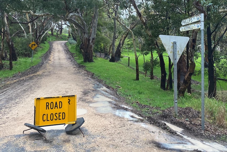 An orange sign reading 'road closed' on a road with gravel and pockets of water