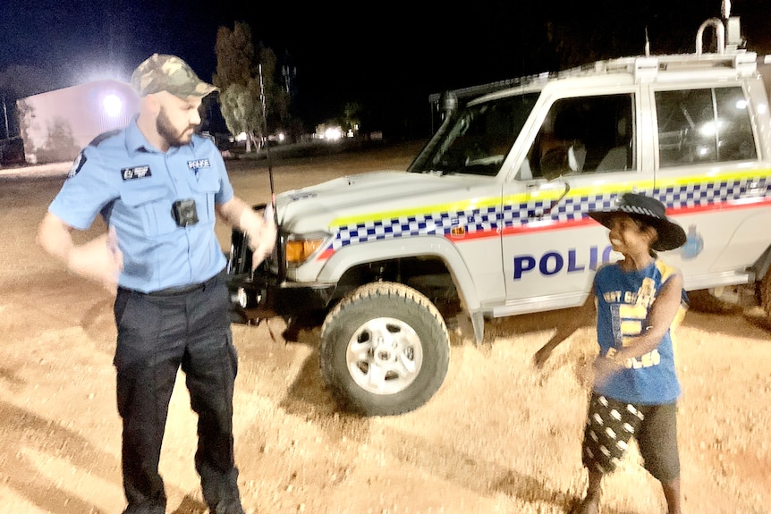 A small child and a uniformed police officer stand laughing in front of a police vehicle