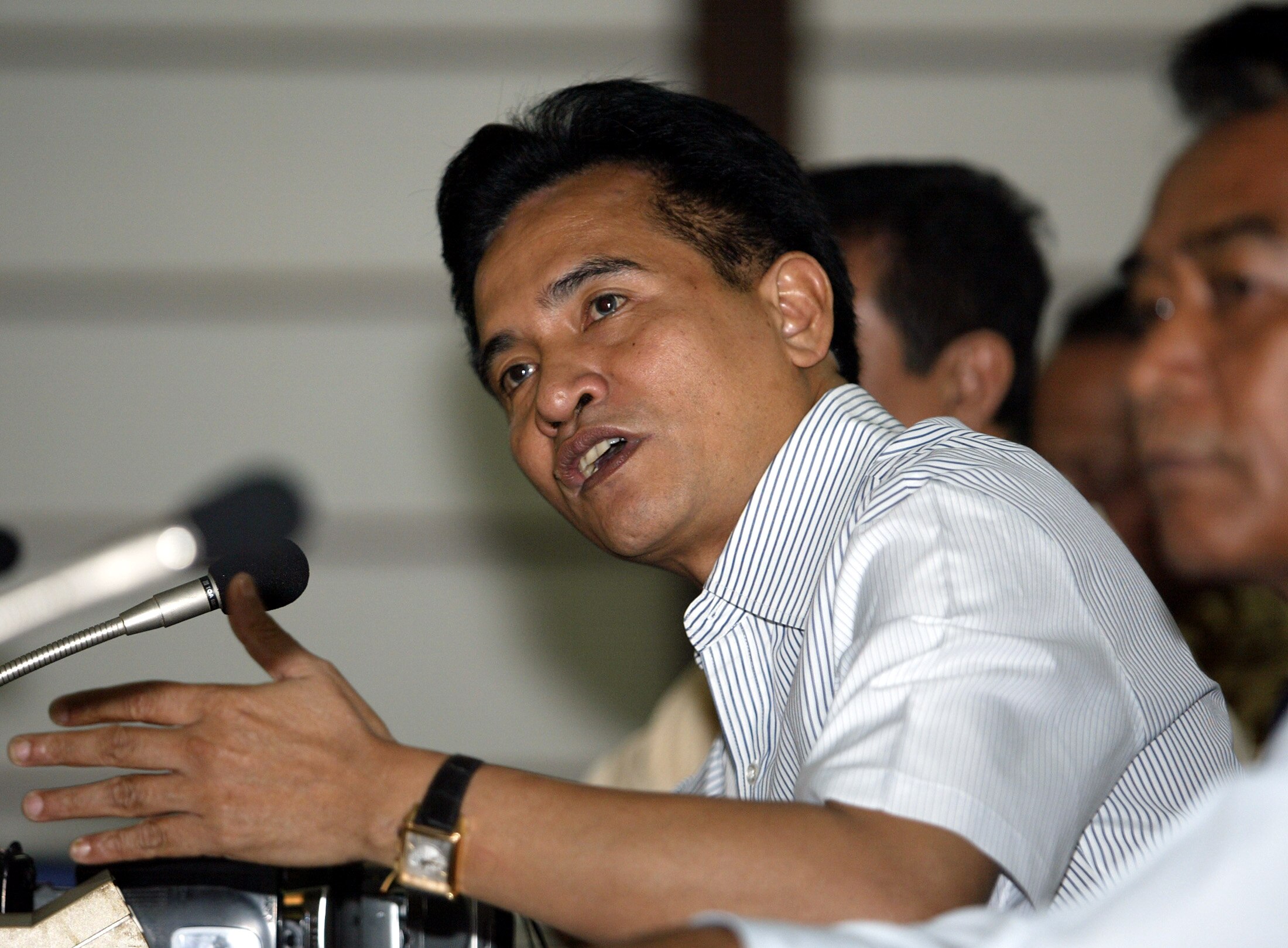 Yusril Ihza Mahendra wearing a white shirt with sleeves leaning over a microphone and gesturing with his left hand