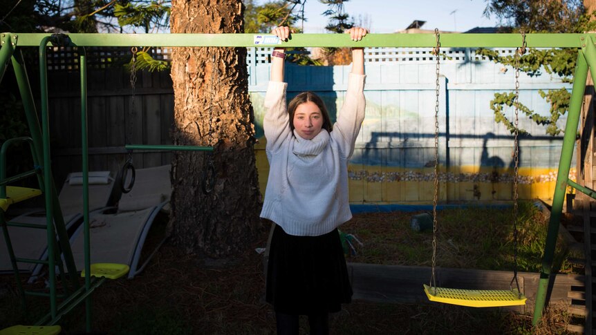 A teenage girl poses for a portrait in late afternoon light, hands rested on a swing set, her shadow cast on the fence behind.