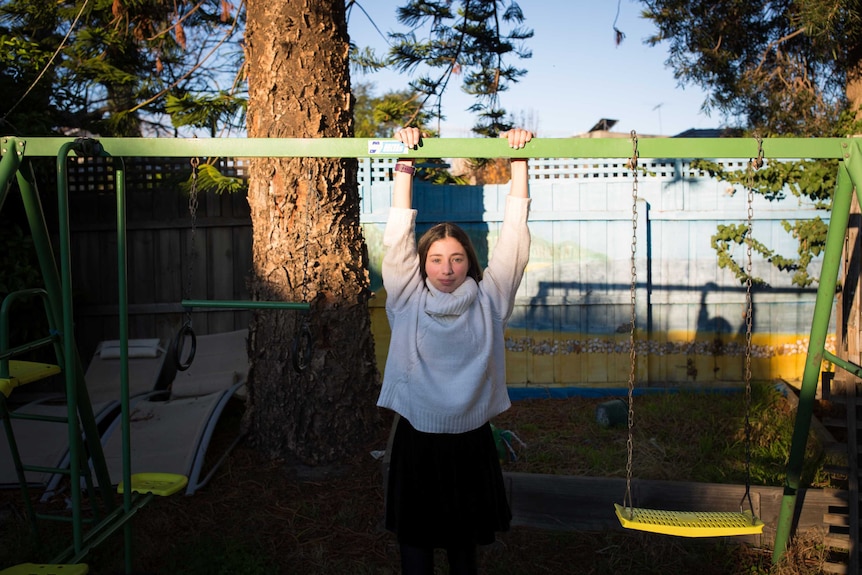 A teenage girl poses for a portrait in late afternoon light, hands rested on a swing set, her shadow cast on the fence behind.
