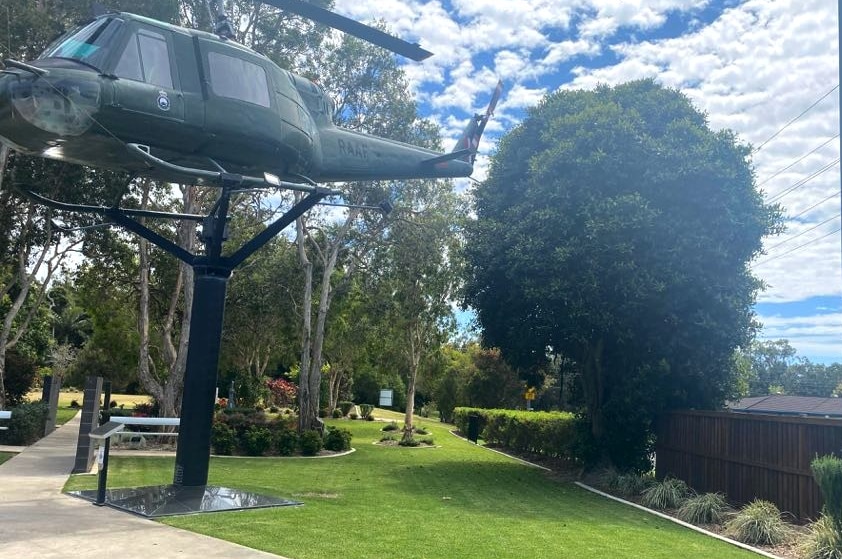 A helicopter mounted on a pole on right with gardens and trees in the background and a garden bed on the right.