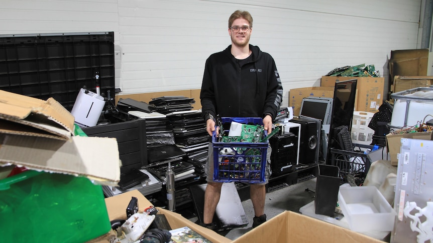 Wade wears a black hoodie and holds a blue milk crate full of electrical goods, surrounded by old computers and cardboard boxes