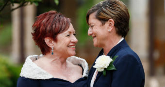 Two women smile at each other lovingly. one wears an off the shoulder dress, the other a navy suit with a flower pinned to chest
