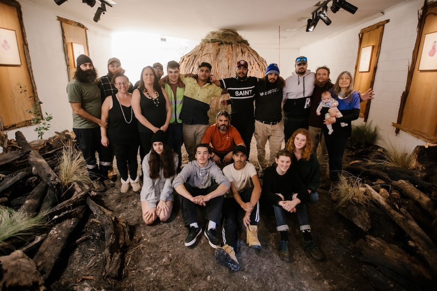 A group of Indigenous and non-Indigenous artists seated or standing in rows in front of a bark hut installation