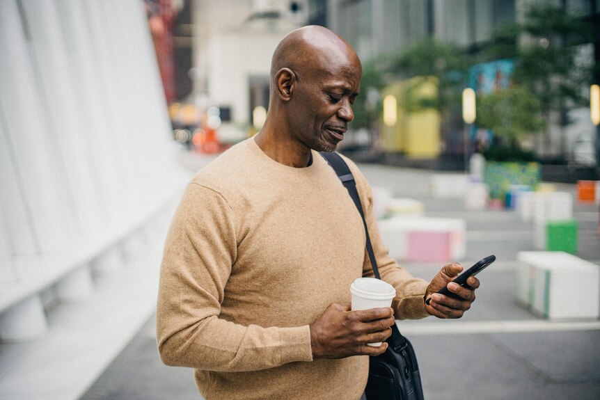 A bald man in a beige sweater holds a take away coffee while using his smartphone.